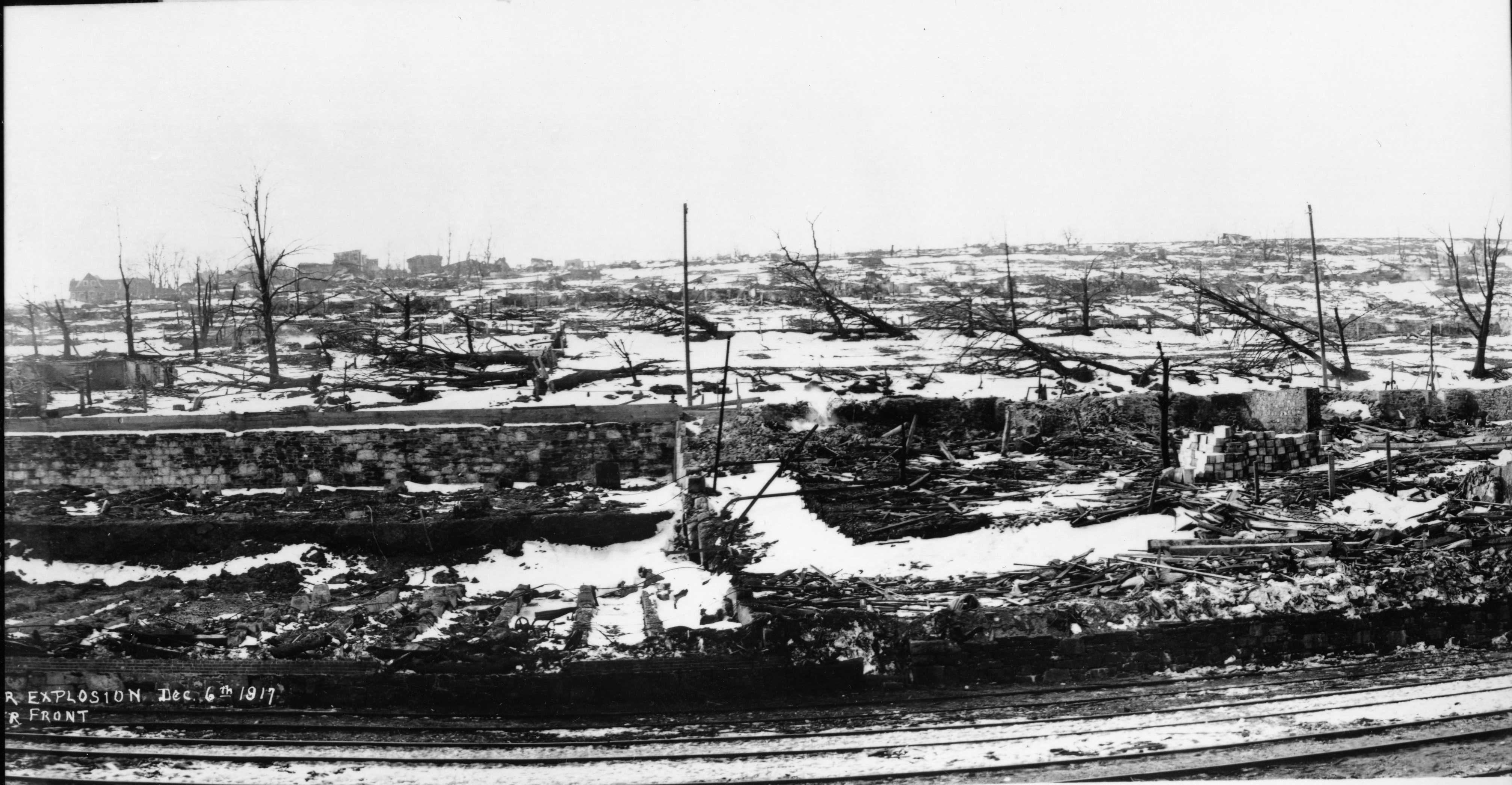 Black and white photograph. Train tracks in immediate foreground. Some structures visible in distance. The foundation and rubble of a building is seen in the centre of the photo. Charred trees and snow on the ground is visible.