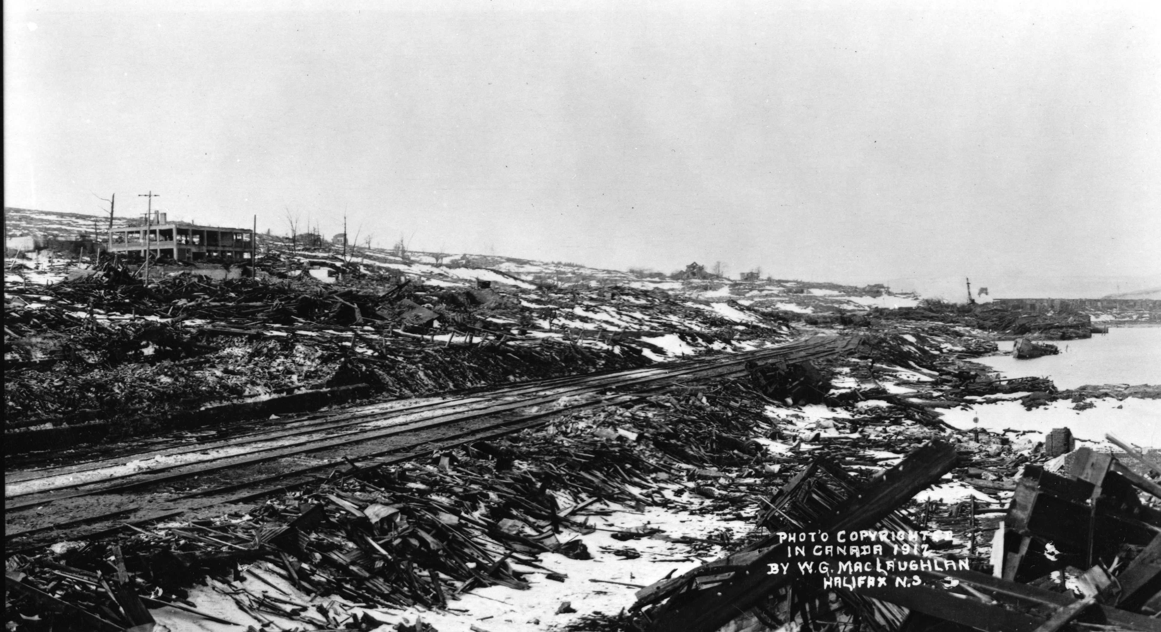 Black and white photograph. Train tracks extend into the distance. A blown out building can be seen; it is still standing but skeletal. Rubble and debris are everywhere. Trees are charred and broken.