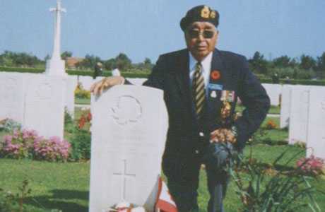 Colour photograph. Mark Wolfleg Sr. is kneeling beside a Commonwealth War Graves headstone, one arm around the top. He wears a Legion blazer and cap.