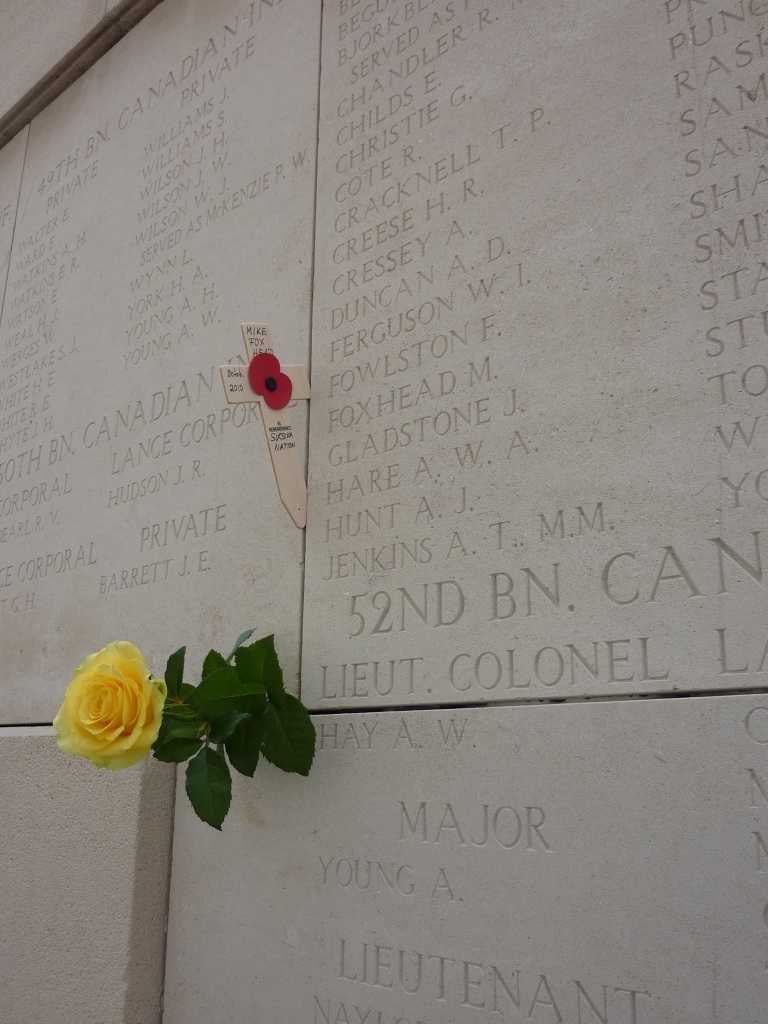 Colour photograph. Among a list of names carved in stone on the Menin Gate, Mike Foxhead is in the centre of the photograph. A wood cross adorned with a poppy and his name and band are stuck in a crack close by; so is a yellow flower.