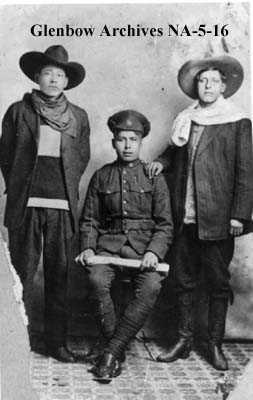 Black and white photograph. Three men are photographed. The men on the left and right are standing, wearing heavy clothing and wide-brimmed hats. In the centre, a young man is seated and dressed in army uniform.