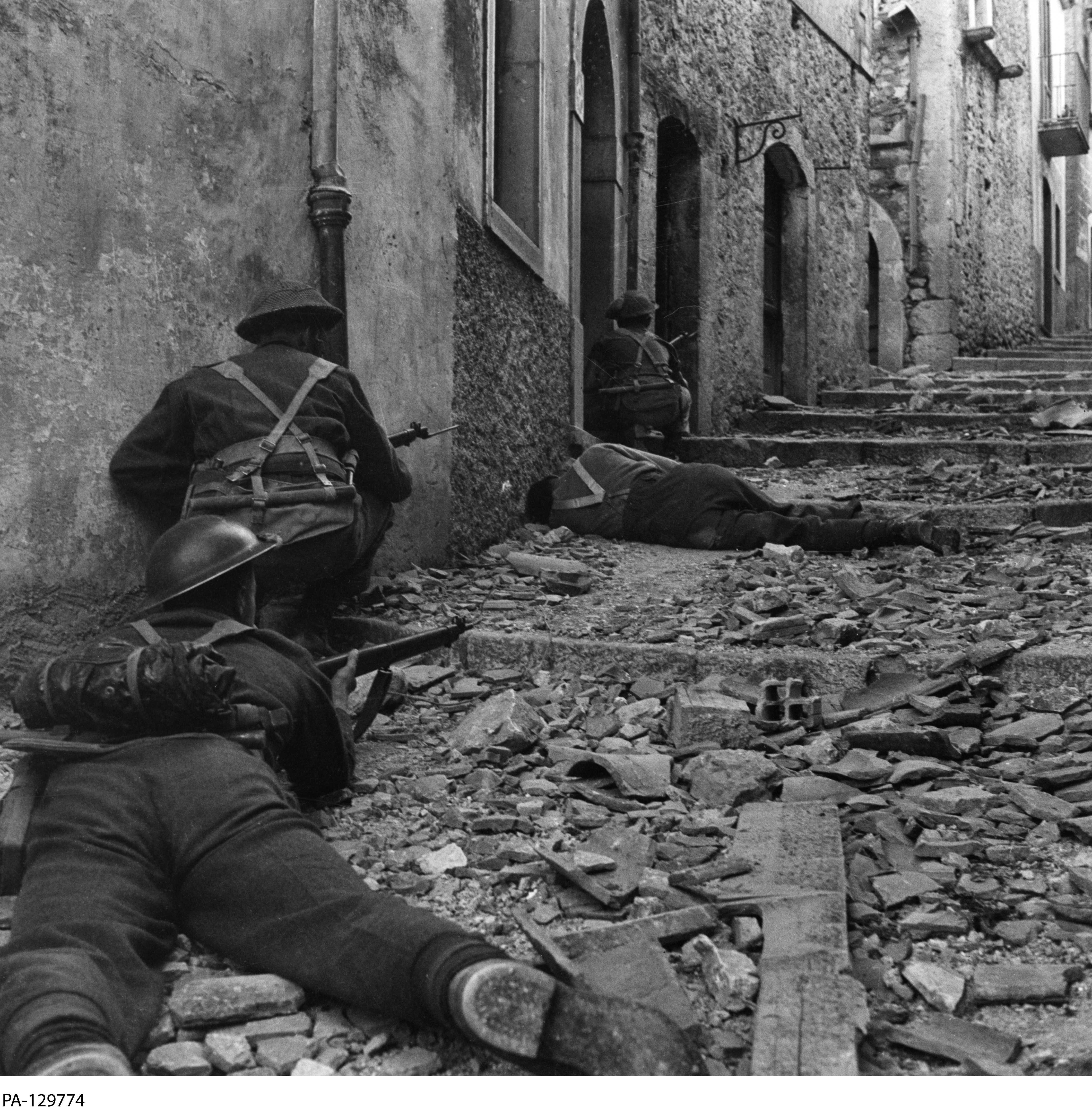 Black and white photograph. Canadian soldiers crouch and aim against a wall on a narrow street. A casualty is visible in front of them.