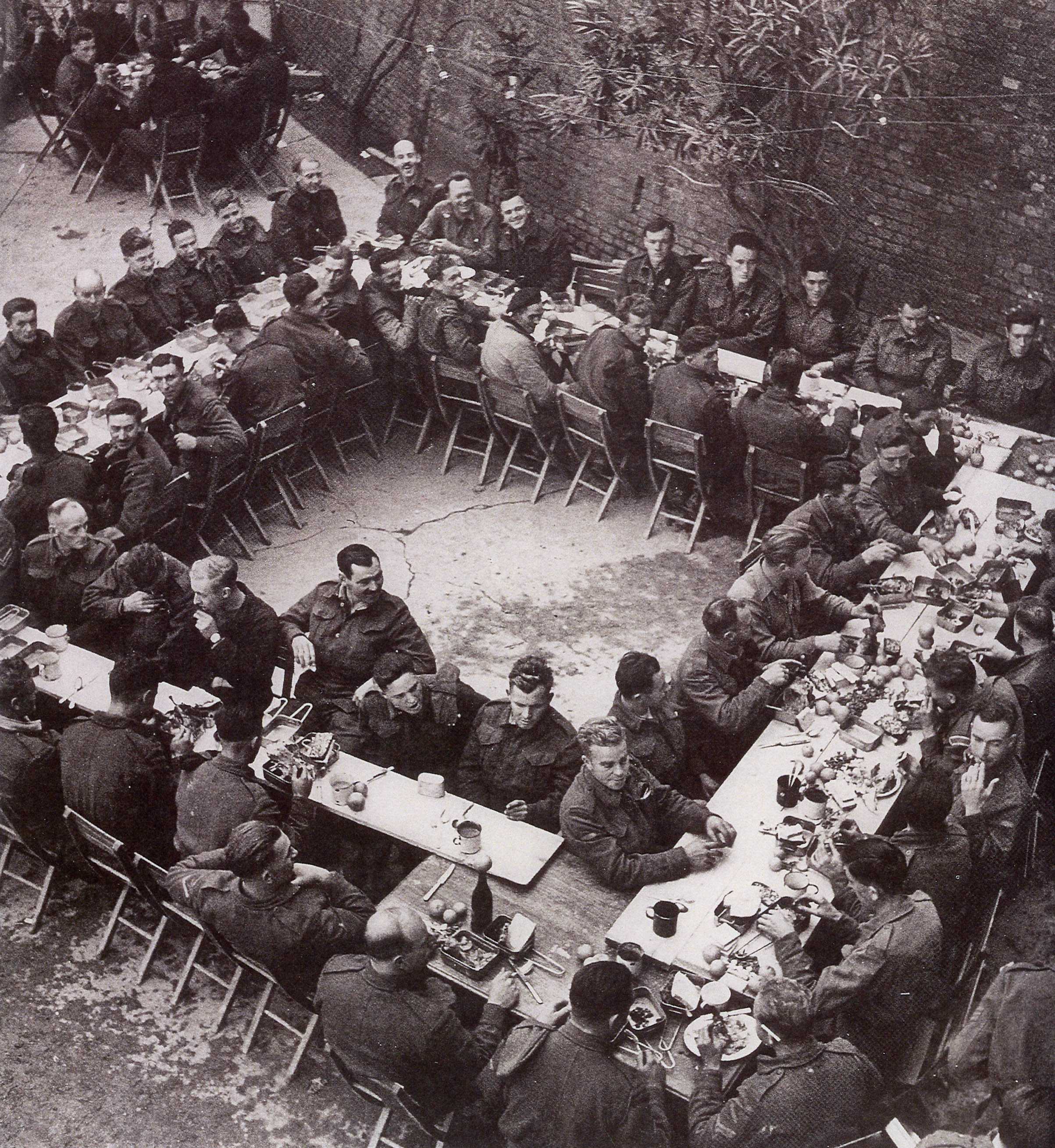 Black and white photograph. Rectangular tables are set up in a large square, viewed from above. Men in military clothing sit on all sides of the tables.
