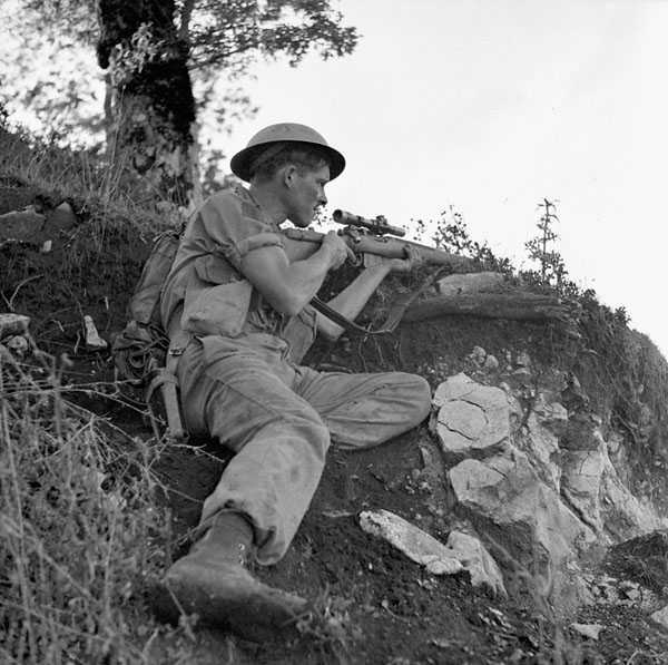Black and white photograph. A Canadian soldier crouches underneath a tree, his body sheltered by a rise in the rocky, brush covered ground. His sleeves are rolled up. He holds  his gun at ready, and his eyes are squinted in concentration.