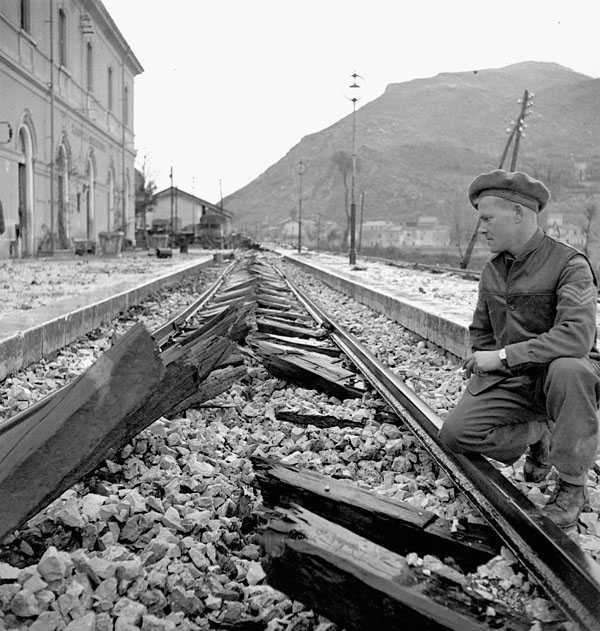 Black and white photograph. A broken rail track runs past a large building and into the mountains. A Canadian kneels beside the rails examining the broken wooden ties, which the camera is focused on. All the ties have been cut in half.