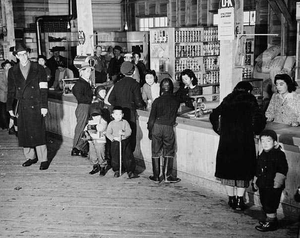 Black and white photograph. People stand in front and behind a store counter, with shelves full of canned food and other stock. People converse across the counter.
