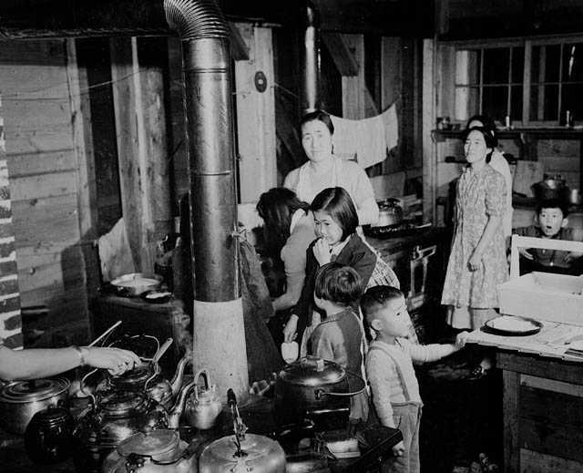 Black and white photograph. Japanese women and children prepare food in a crowded kitchen. A stove covered in kettles is in the foreground, towels hung on a line are visible in the background.