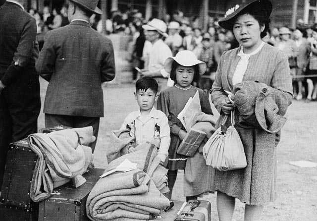 Black and white photograph. A women stands with two children, dressed in their Sunday best. Bundles of possessions and suitcases sit nearby.