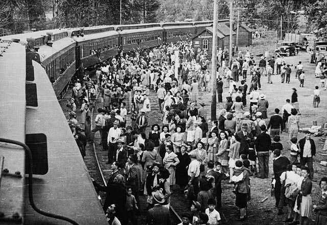 Black and white photograph. A passenger train at a station. A large crowd of stands alongside, some people talk to those already on board the train.