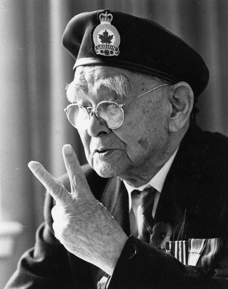 Black and white photograph. An elderly Masumi Mitsui gives a peace sign. He wears his Legion blazer, medals, and a poppy.