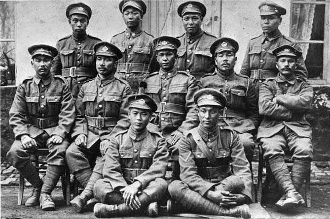 Black and white photograph. The image shows eleven Japanese-Canadian soldiers dressed in First World War Canadian military uniforms and hats, seated and standing in three rows outside of a light-coloured building.