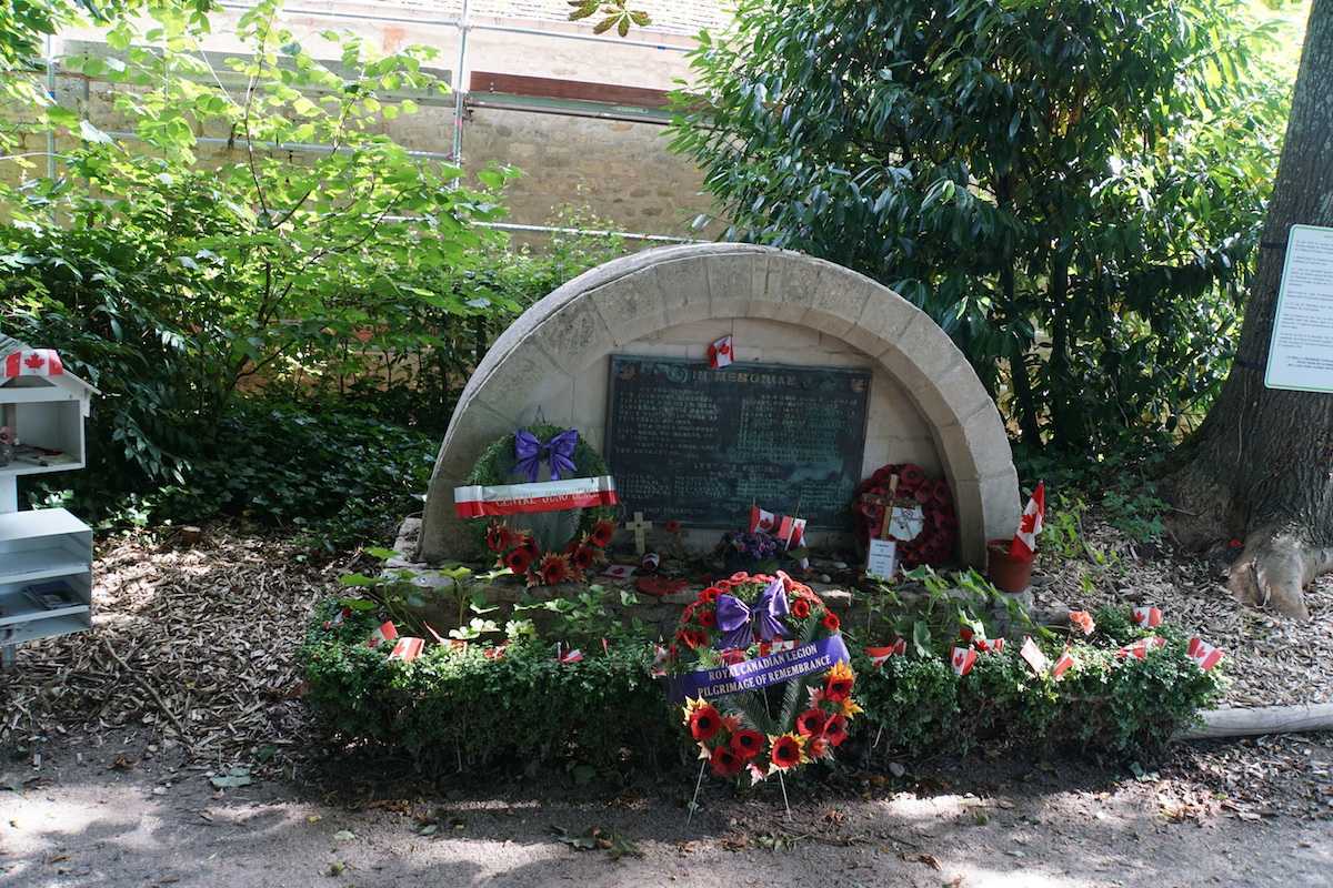 Colour photograph. A small, half circle stone memorial sits amidst a lush garden. Several commemorative wreaths are laid at its base.