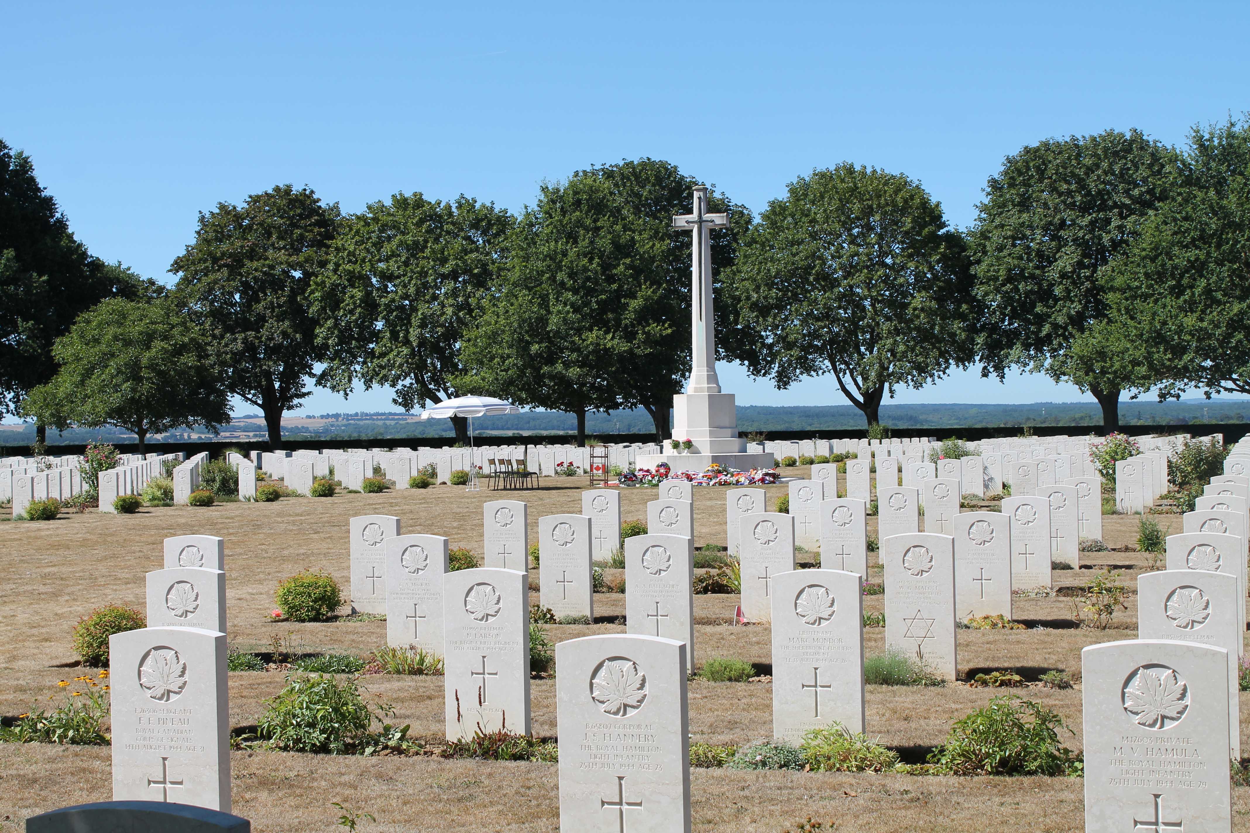 Colour photograph. A view of a military cemetery with uniform white gravestones. The iconic Commonwealth War Graves marble cross is in the centre. A podium from an earlier ceremony remains.