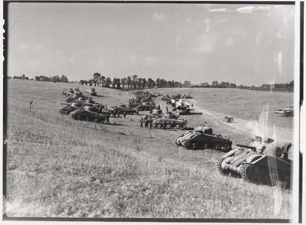 Black and white photograph. Along a road through a file which approaches a forested area, a large concentration of tanks sit on the sloped area to the left of the road. Military personnel mingle around the tanks.