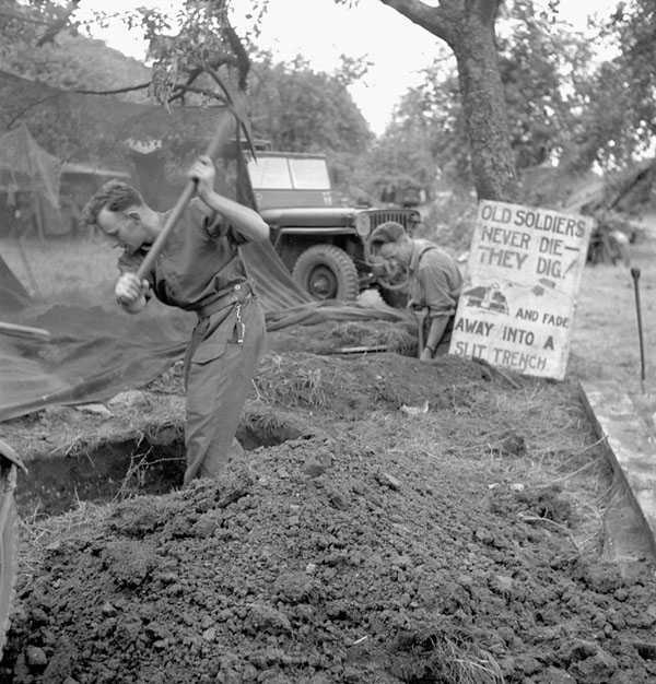 Black and white photograph. Two men dig a narrow trench in the ground. Tarps and a military jeep are in the background, in front of a forested area.