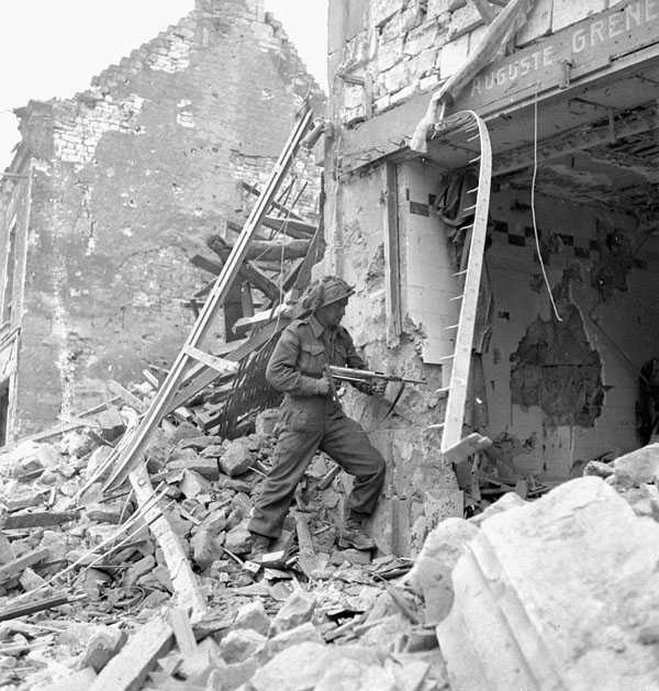 Black and white photograph. A soldier stands on a large pile of rubble pointing a gun into a gaping hole in the front of a building.  Another ruined building is visible behind him, and the street is entirely covered in rubble.