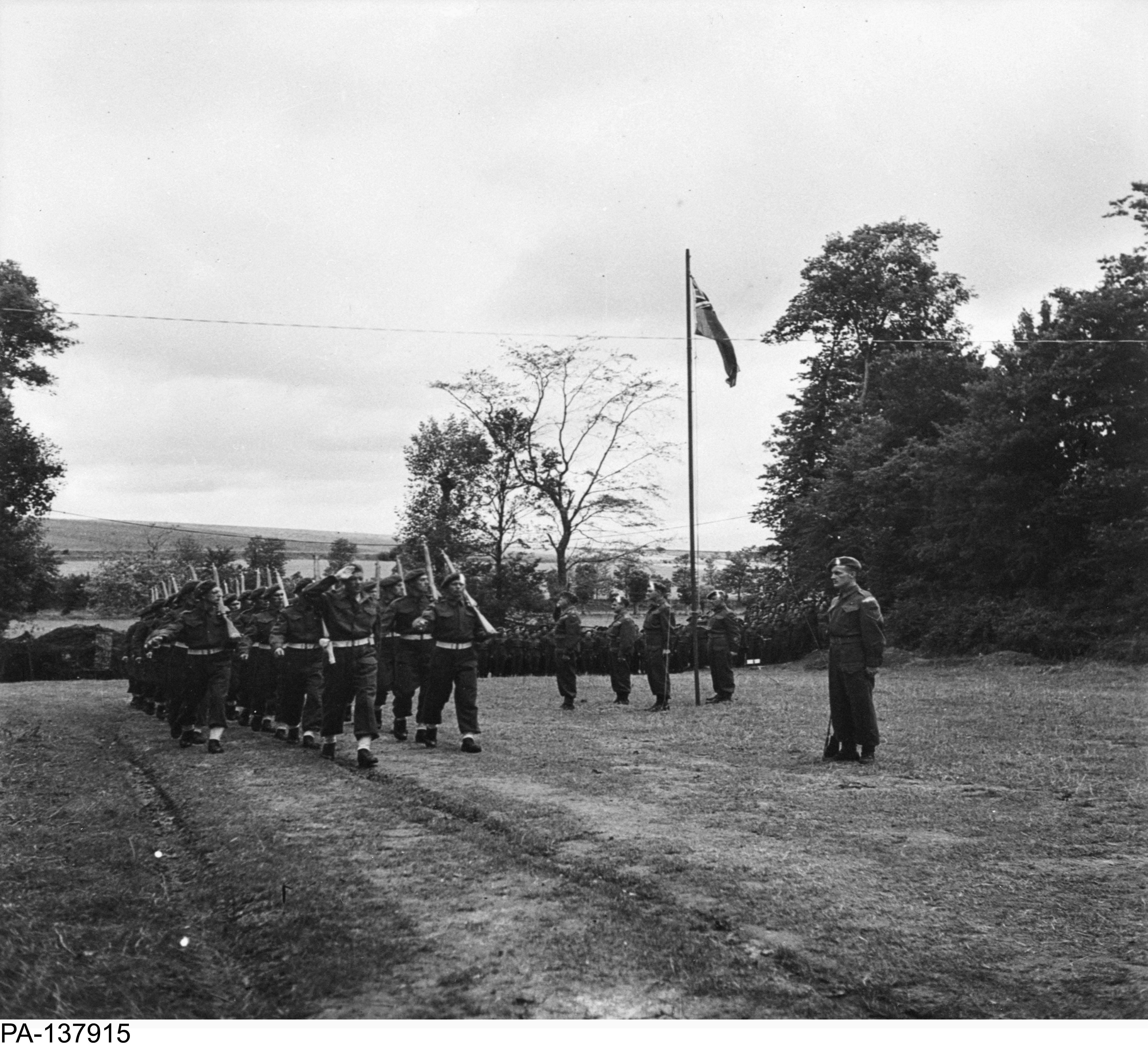 Black and white photograph. Three rows of soldiers march past a raised red ensign and superior officers. All men salute. They are outside, marching across a grassy field surrounded by trees.