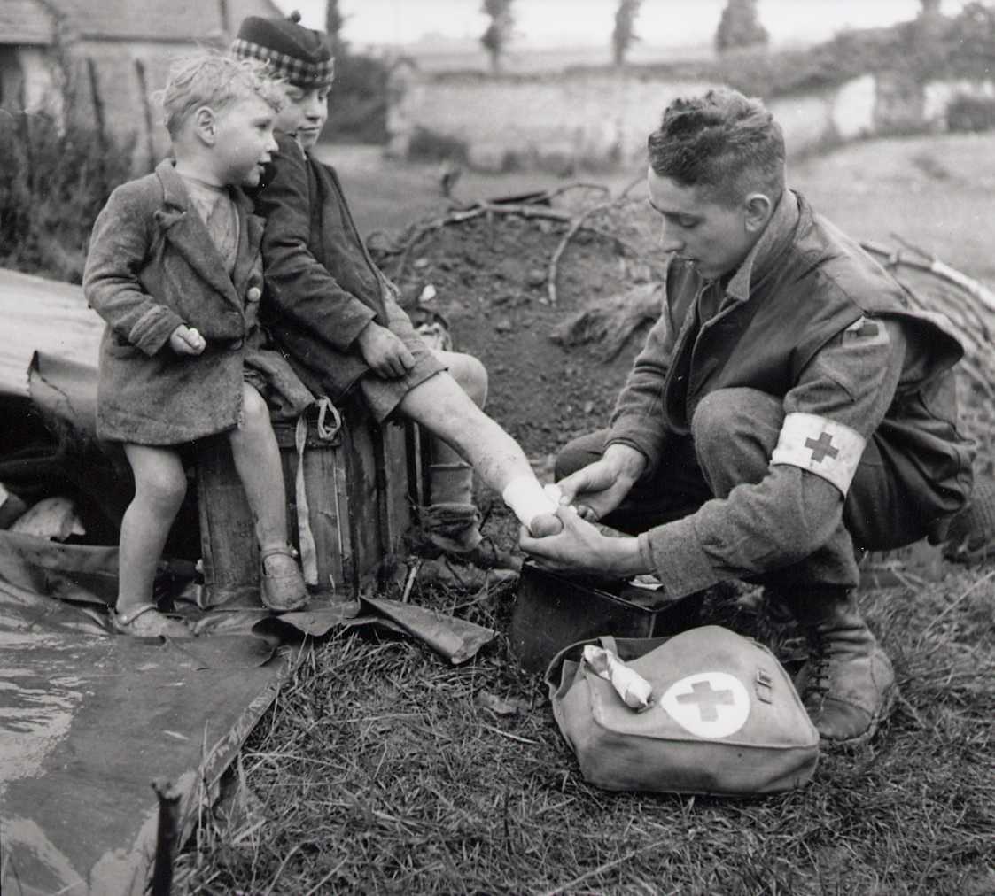 Black and white photograph. A soldier (with medical arm band) bandages the ankle of a young boy while another young child watches.