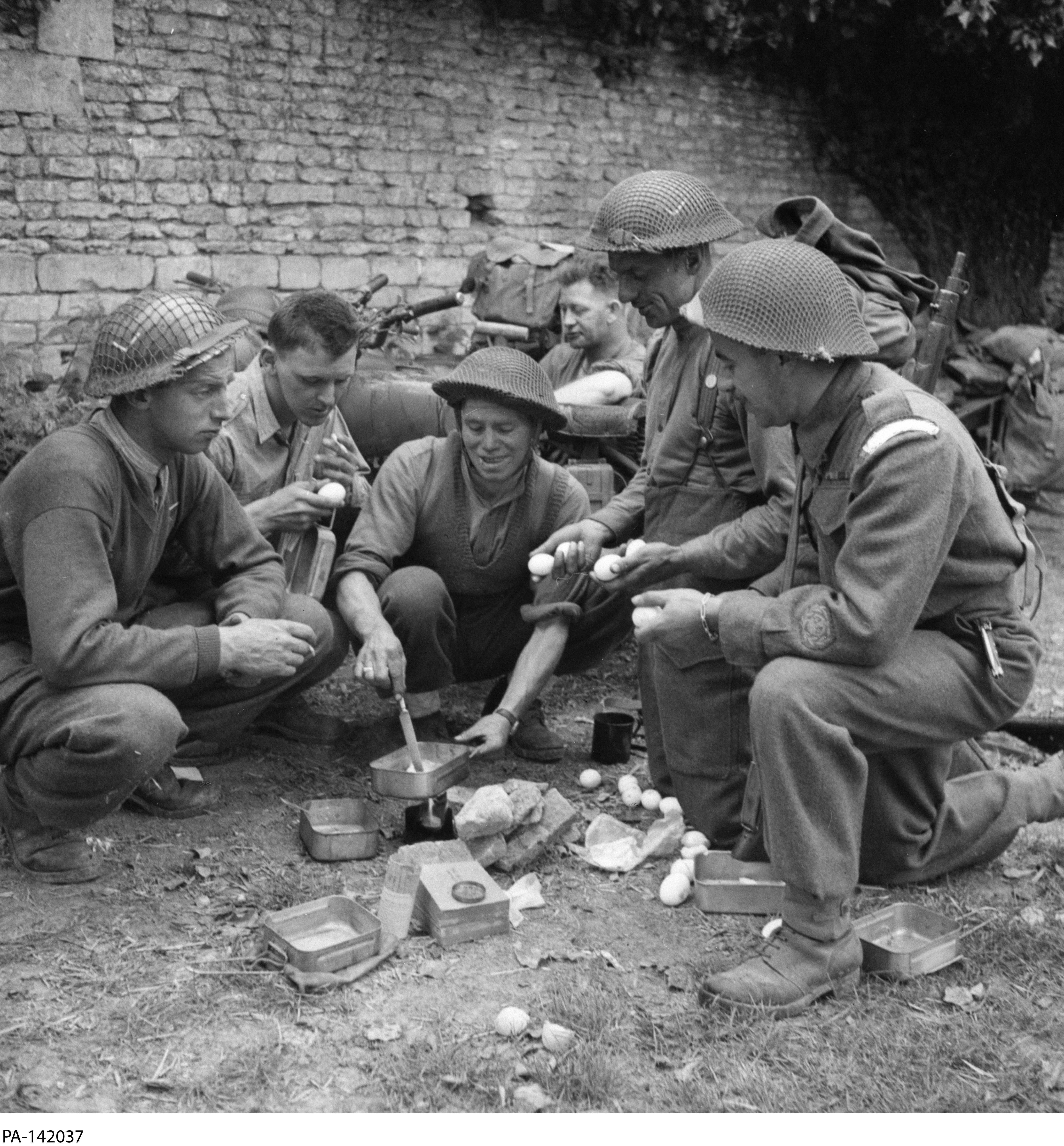 Black and white photograph. Five soldiers crouch around a small flame. One holds a mess tin over the flame, stirring something inside. There are eggs on the ground, waiting to be cooked. Other soldiers mingle behind them.