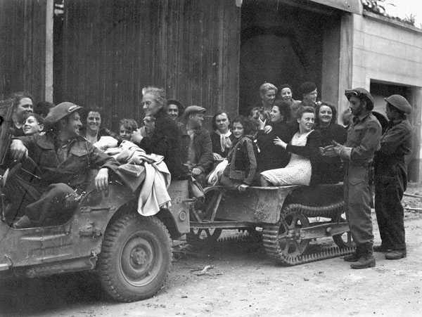 Black and white photograph. A military jeep and trailer are loaded with French civilians. A Canadian soldier is driving and a few others stand alongside.