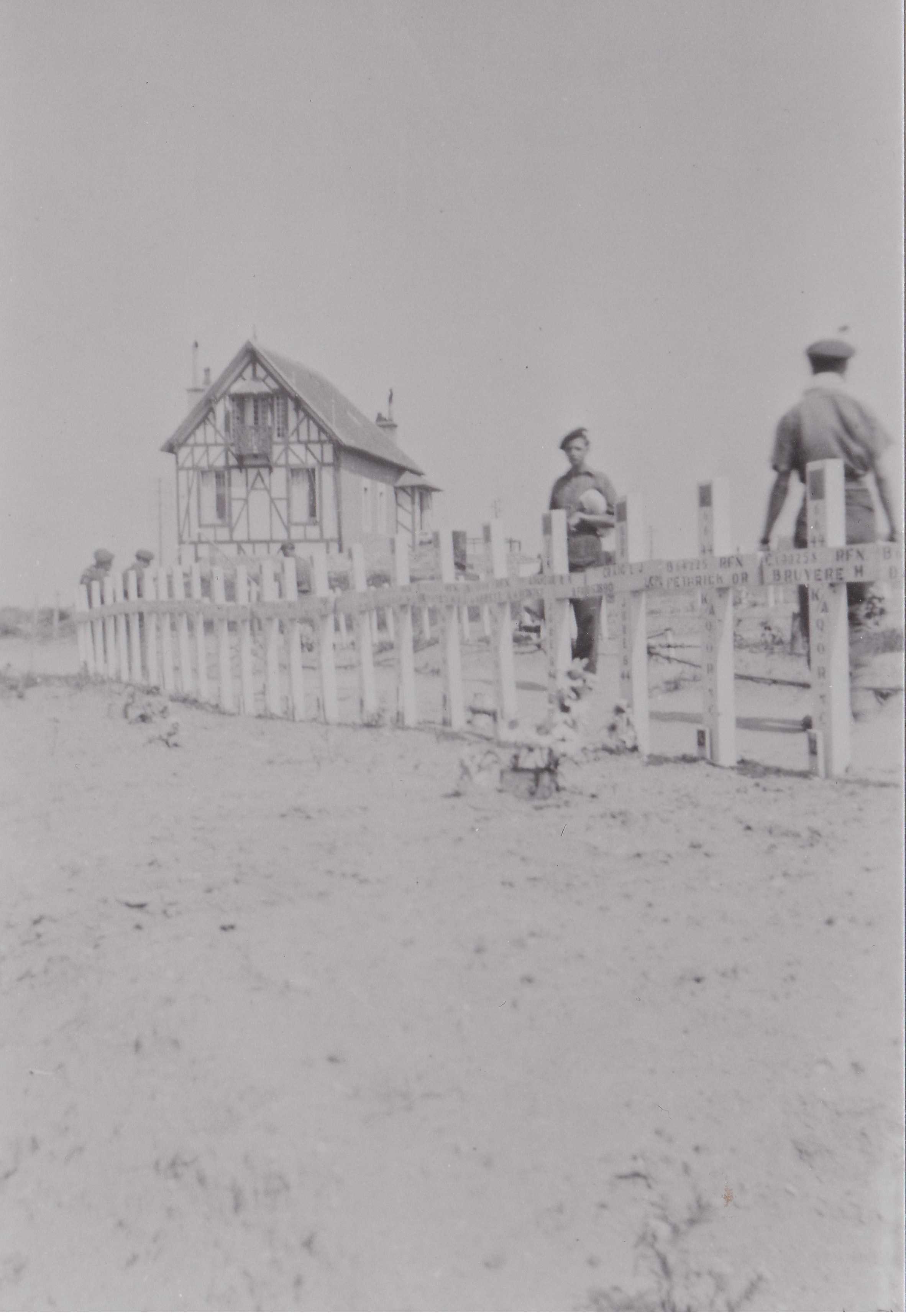 Black and white photograph. With a typical French home in the background, two men walk behind a row of white crosses planted in the ground. The photo is washed out, but it is clear that there are names on every cross.