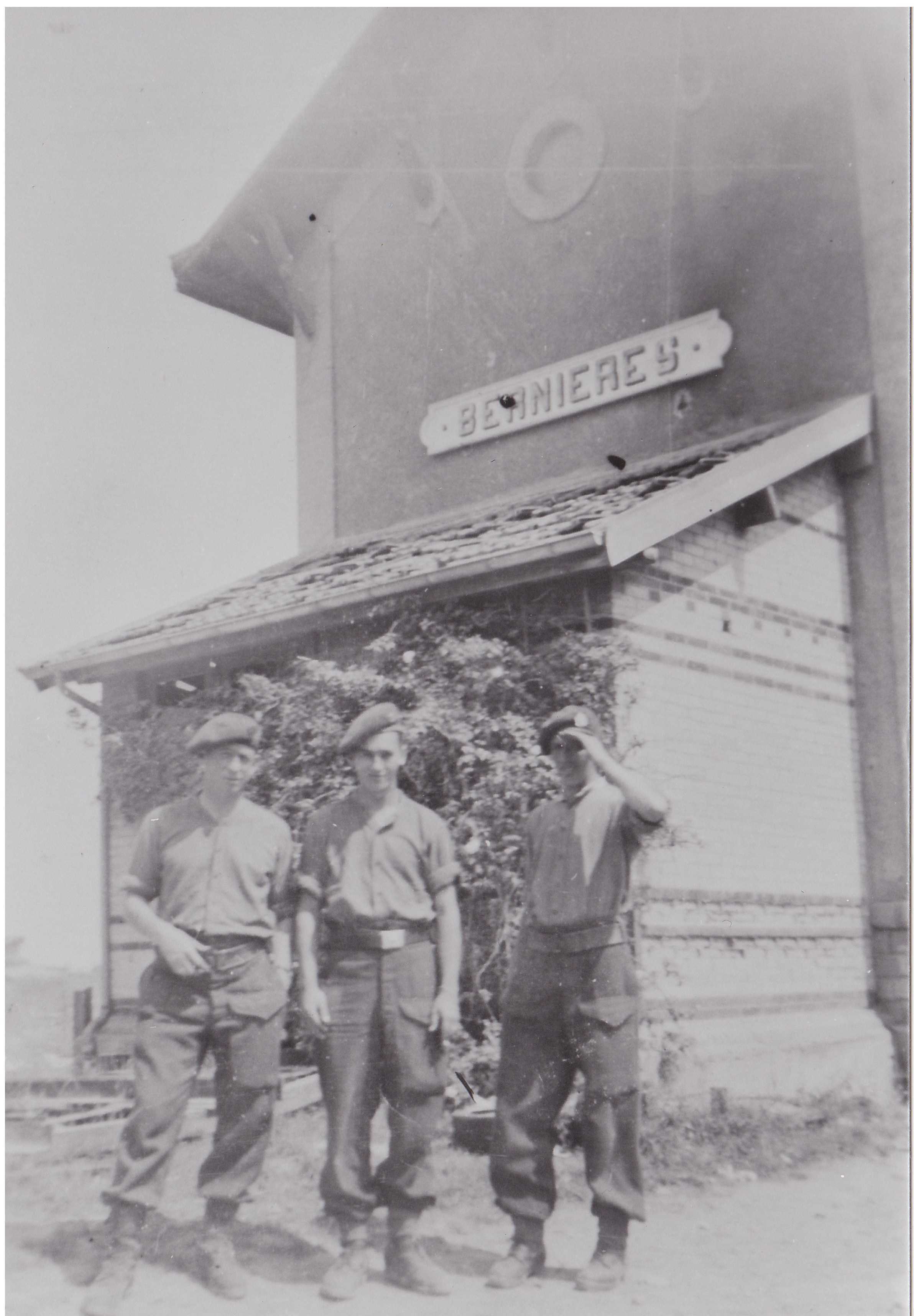 Black and white photograph. Three Canadian men in casual military uniforms stand in front of a large building labelled "Bernieres." One holds his cap down over his eyes to shield the sun.