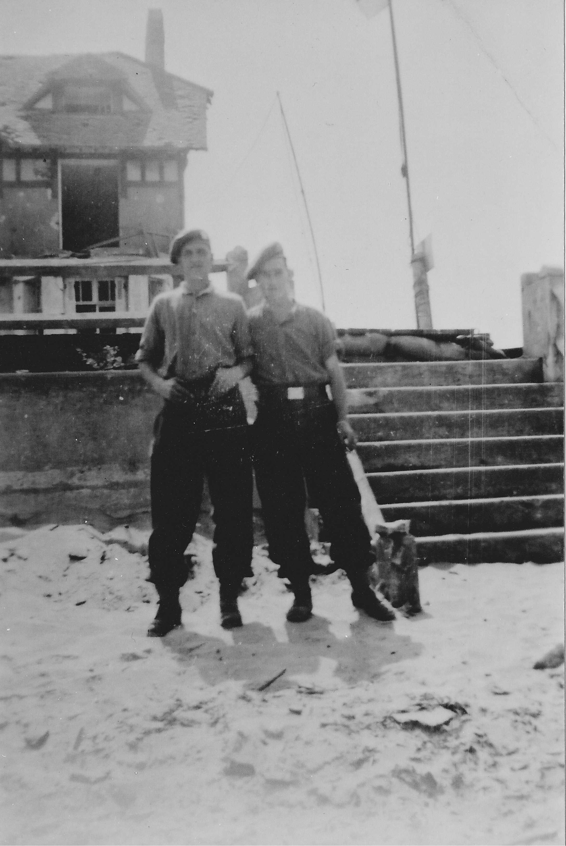 Black and white photograph. Two Canadian soldiers stand in front of the sea wall; there is a stone staircase to their left. A house is visible in the background, with a window blown out. They stand on a sandy beach.
