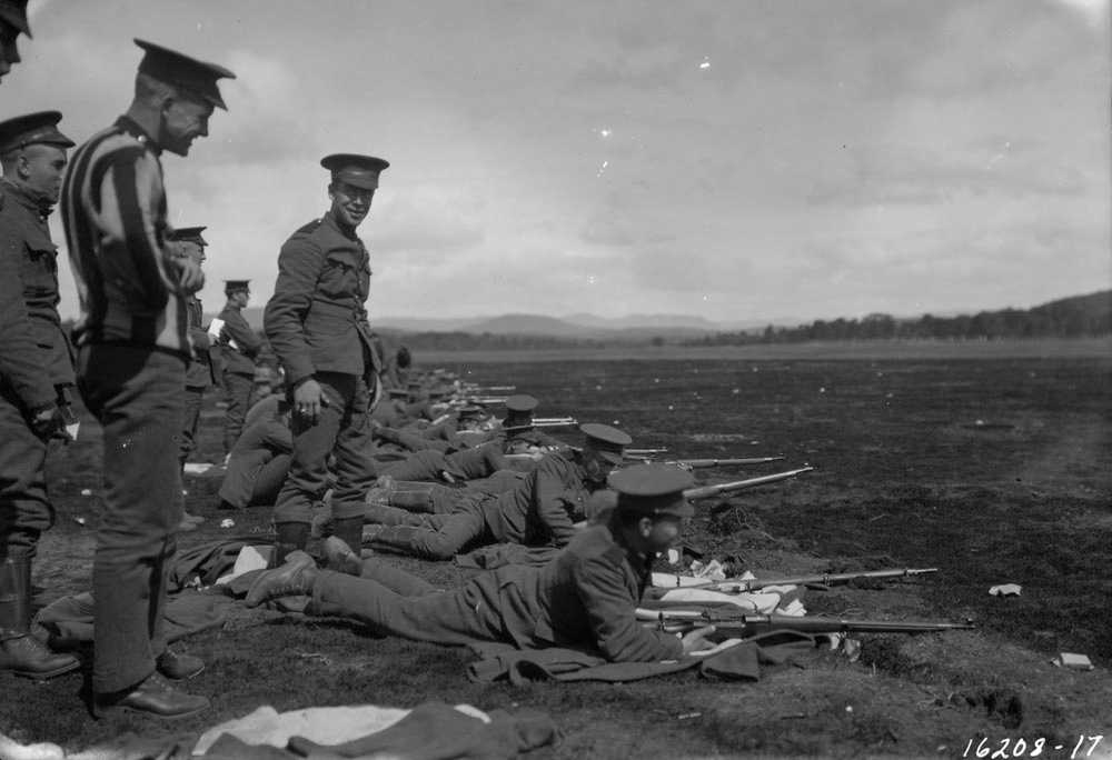 Black and white photograph. In a large field, men lie on the ground learning to aim Ross rifles; trainers stand above them. All are outfitted with military uniforms.