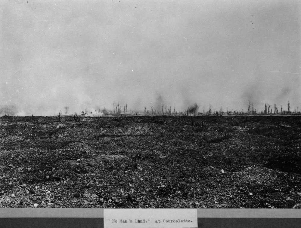 Black and white photograph. Shells can be seen exploding in the distance over barren No-mans land (labelled as such at the bottom of the image). The ground is muddy and uneven, dead trees are scattered.