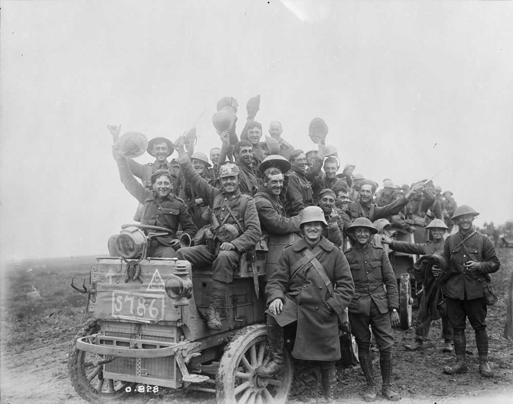 Black and white photograph. A large group of Canadian soldiers hang off or stand beside military vehicles. They raise their hats in salute to their victory, and look generally cheery.