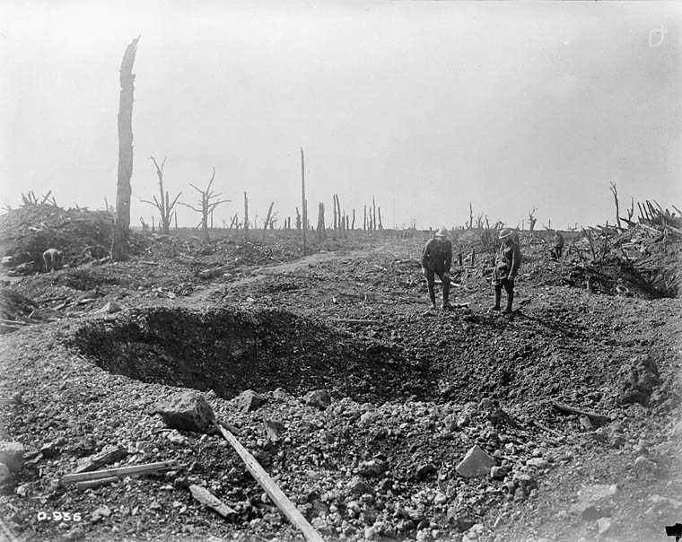 Black and white photograph. A large shell hole in the middle of the road. Two men in military uniforms and helmets stand behind it, one crouching slightly. The landscape is rough and rocky.