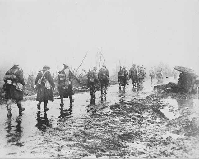 Black and white photograph. A group of soldiers walk away from the camera single file. The road is covered in mud and water.  The soldiers carry packs on their backs.