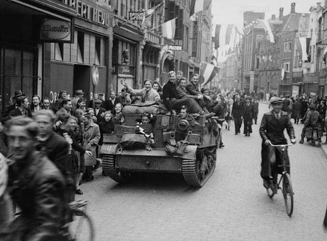 Black and white photograph. Street scene. Dutch flags are everywhere, in hands and hanging. People crowd the sidewalks and streets on foot and bike. A tank drives down the centre of the street, and civilians run alongside and hang off of it.