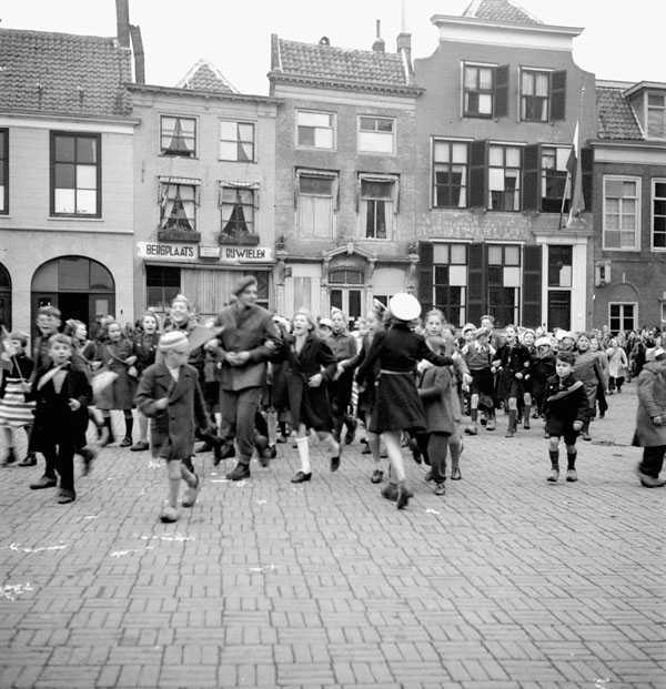 Black and white photograph. Cobblestoned town square, narrow building in background. A large group of children in warm coats circulate and move through the square, looking generally excited. Many wave small dutch flags. There is a Dutch flag hanging on a