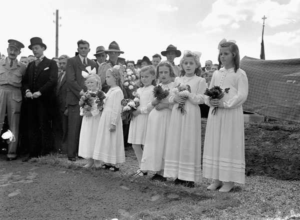 Black and white photograph. 6 young Dutch girls stand in a row, wearing white dresses and holding bouquets of flowers. Onlookers in civilian and military clothing stand behind them.