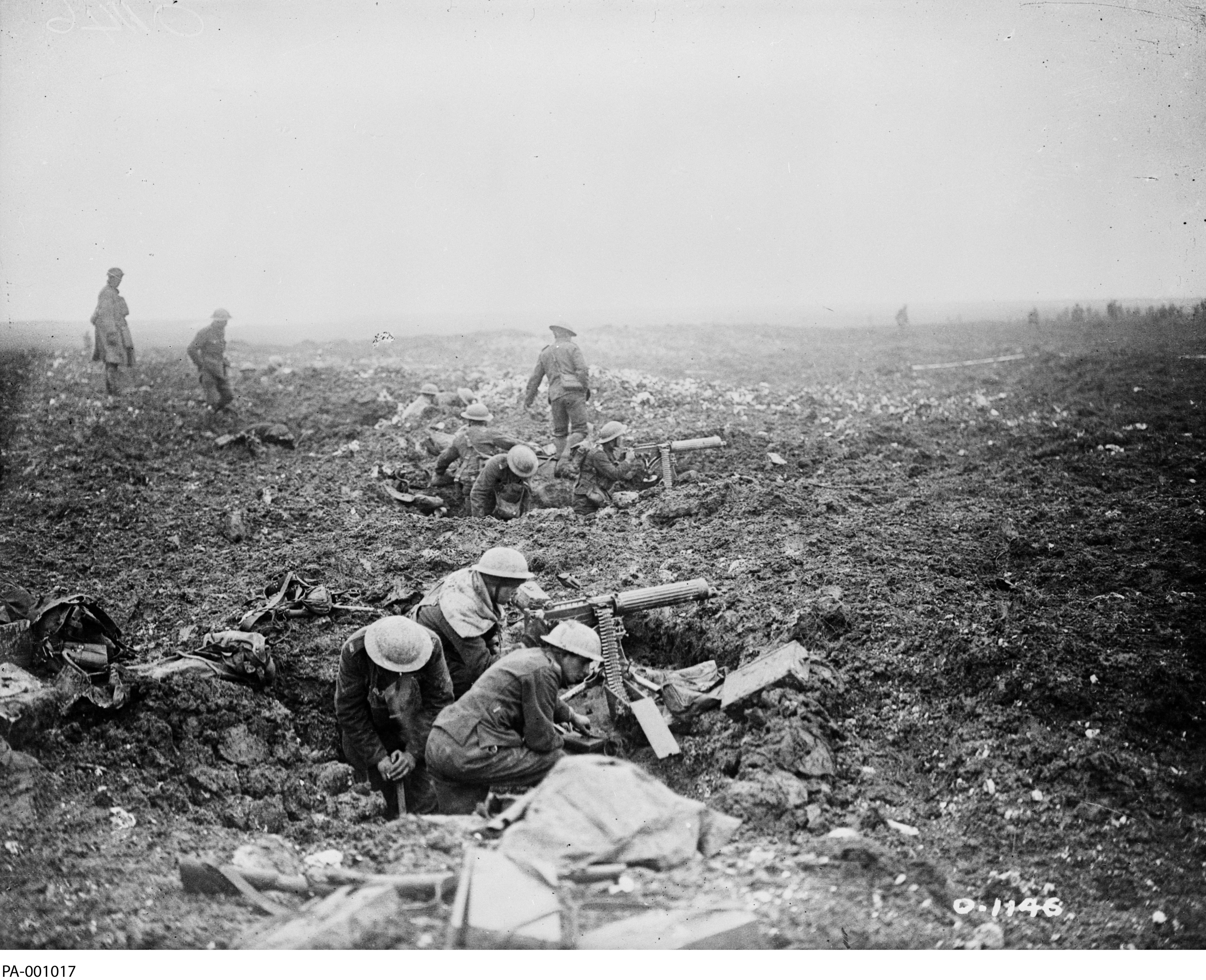 Black and white photograph. Two large holes in the ground. In each one, 3-4 men in military gear arm and aim a machine gun. The ground is muddy and the air is filled with smoke. Several men walk behind them.