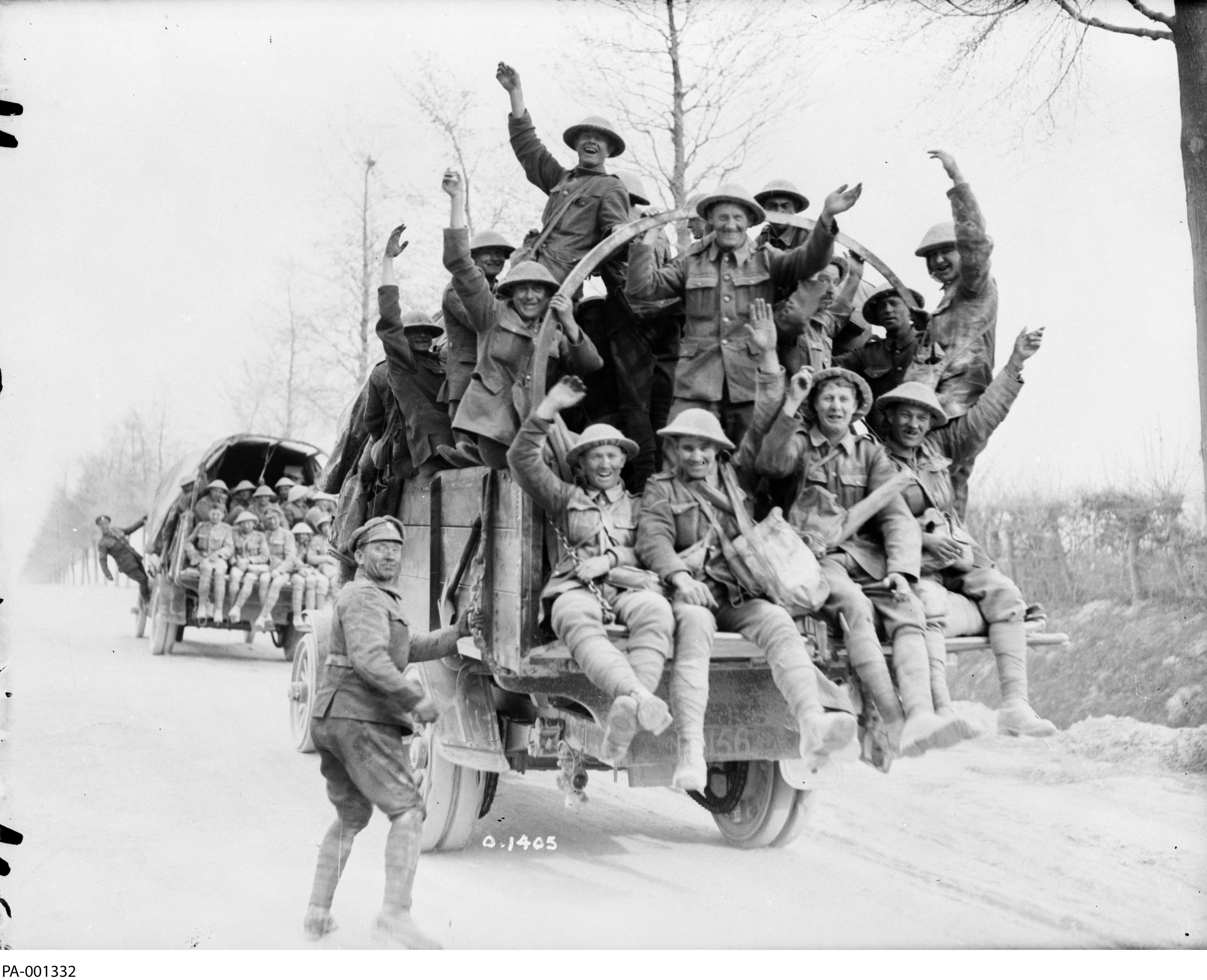 Black and white photograph. Canadian soldiers face the camera; they are all seated on the back of the truck, one soldier is just about to climb on. They appear happy and are waving. Another truck is visible in the distance, similarly loaded with soldiers.