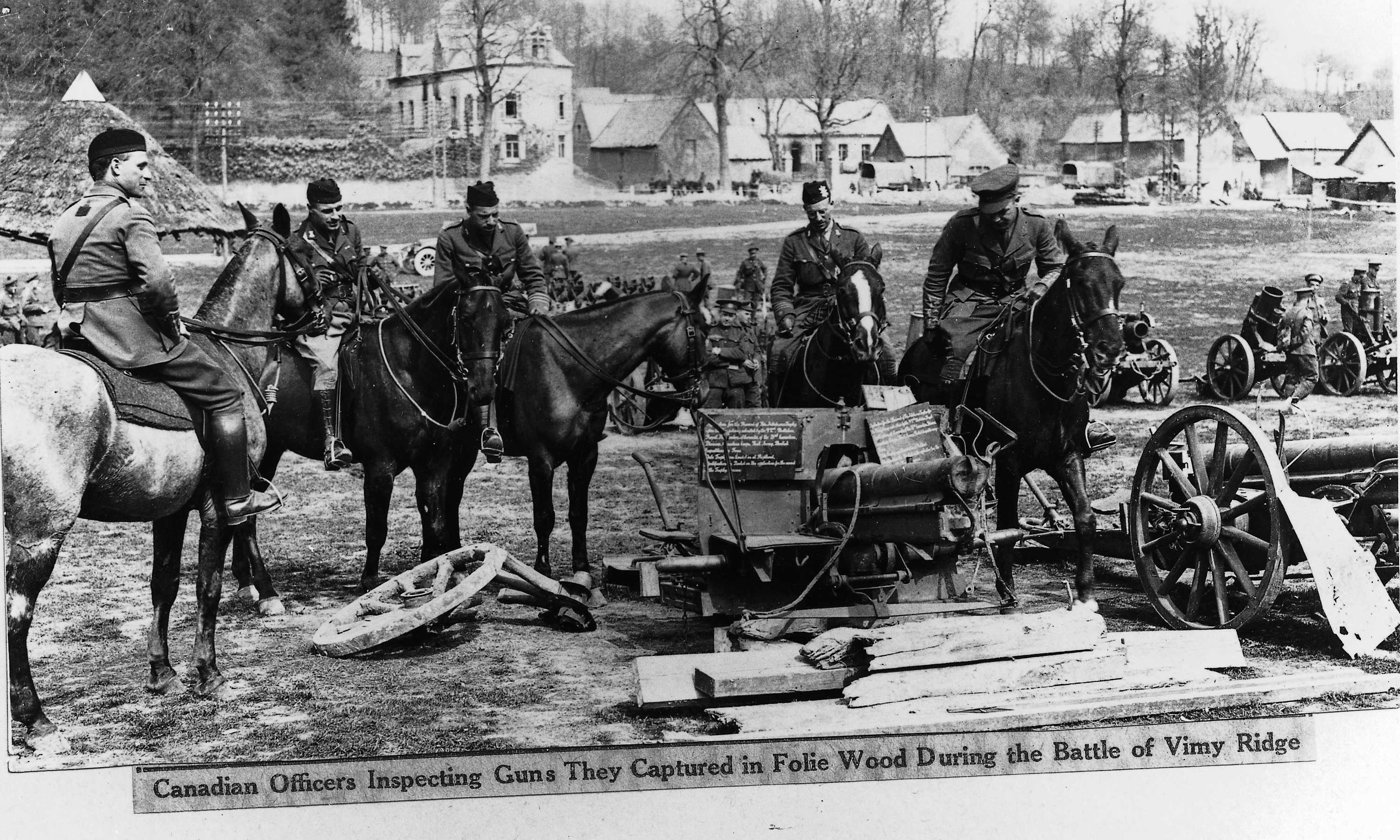 Black and white photograph. Men in military dress and sitting on horses look down over a pile of guns mounted on wheels for easy movement. There are piles of large pieces of wood on the ground, and a village visible in the distance behind them.