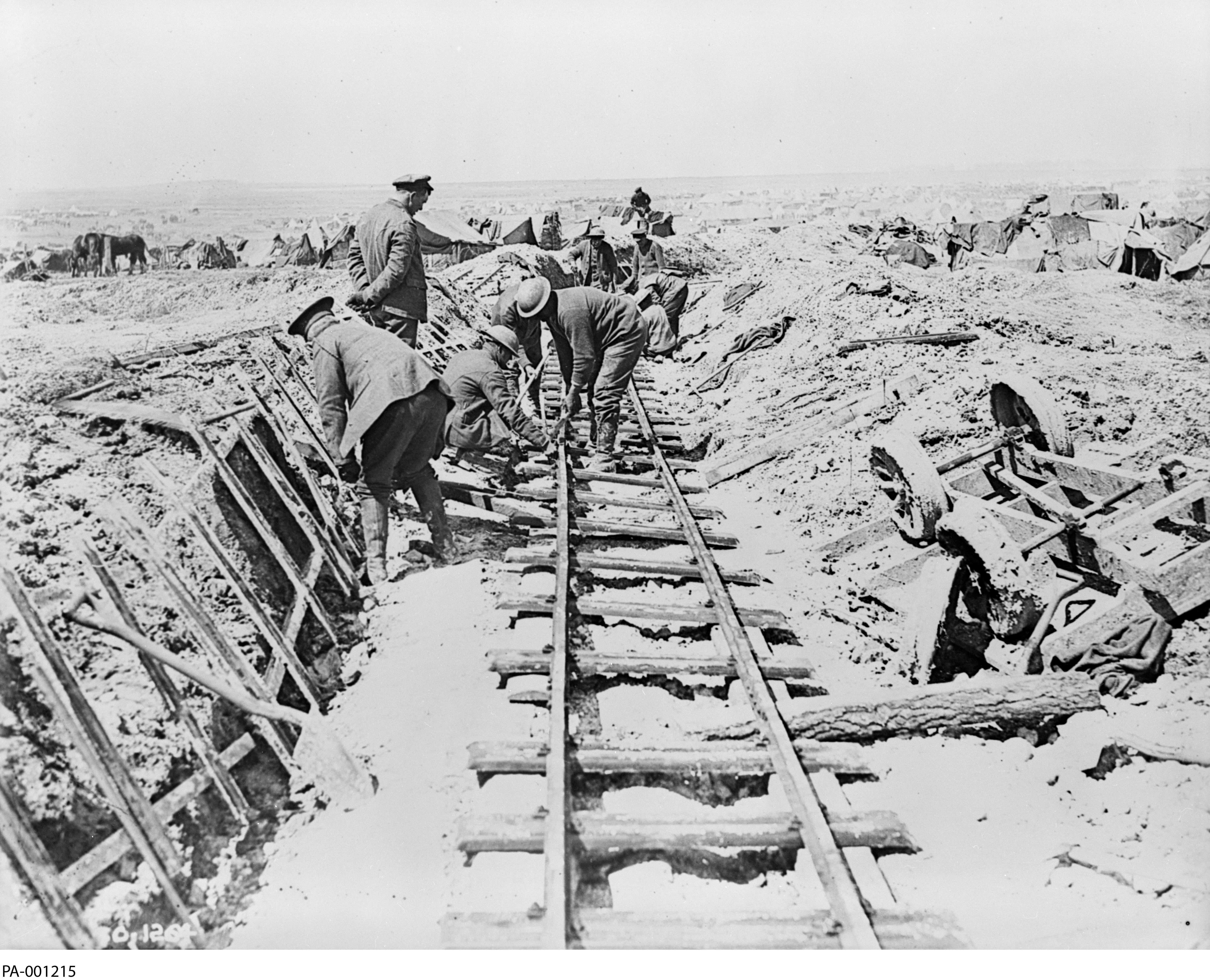 Black and white photograph. In the foreground, on muddy ground, 4 men lay pre-assembled rail tracks. A 5th man, dressed in civilian clothing, watches.